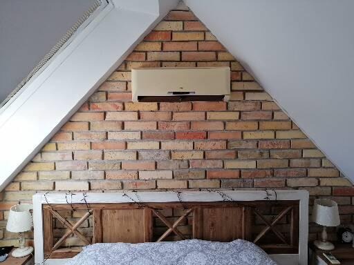 Klimatyzator zamontowany nad wezgłowiem łóżka na ścianie z cegły w sypialni na poddaszu.