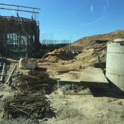 Plac budowy z widoczną konstrukcją z rusztowań, stertą zbrojeń, betonowymi kręgami i ziemnym nasypem w tle, całość skąpana w jasnym świetle słonecznym.