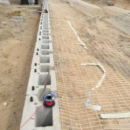 Wznoszenie betonowej ściany oporowej z pustaków szalunkowych na gruncie wzmocnionym geokratą, widoczny zwój czerwonego drutu do wiązania zbrojenia oraz młotek murarski.