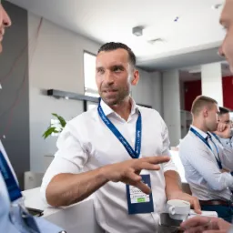 Mężczyzna w białej koszuli gestykuluje podczas rozmowy z dwoma innymi mężczyznami, trzymając filiżankę kawy i identyfikator z napisem 'Kapitał'.