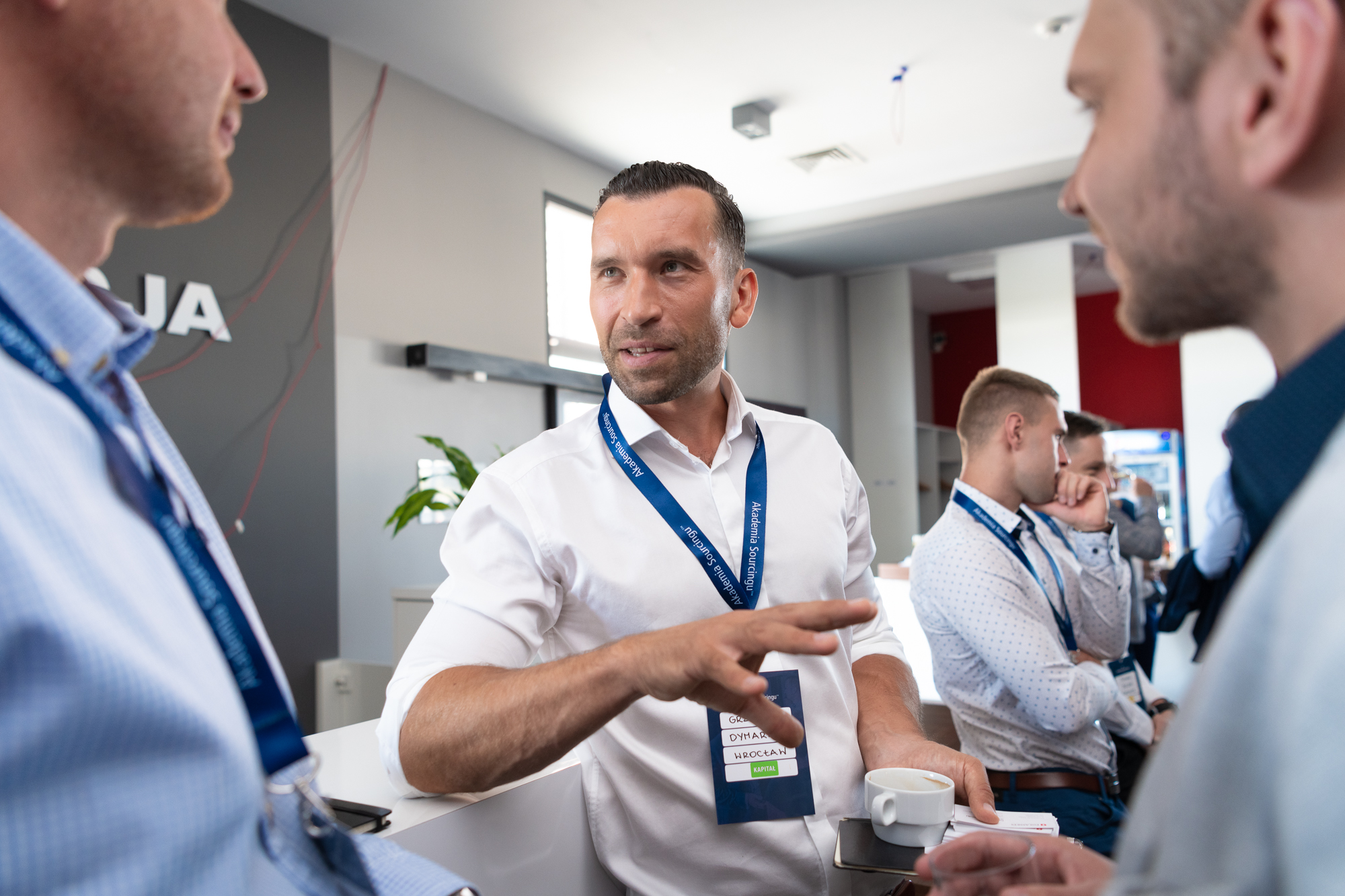 Mężczyzna w białej koszuli gestykuluje podczas rozmowy z dwoma innymi mężczyznami, trzymając filiżankę kawy i identyfikator z napisem 'Kapitał'.