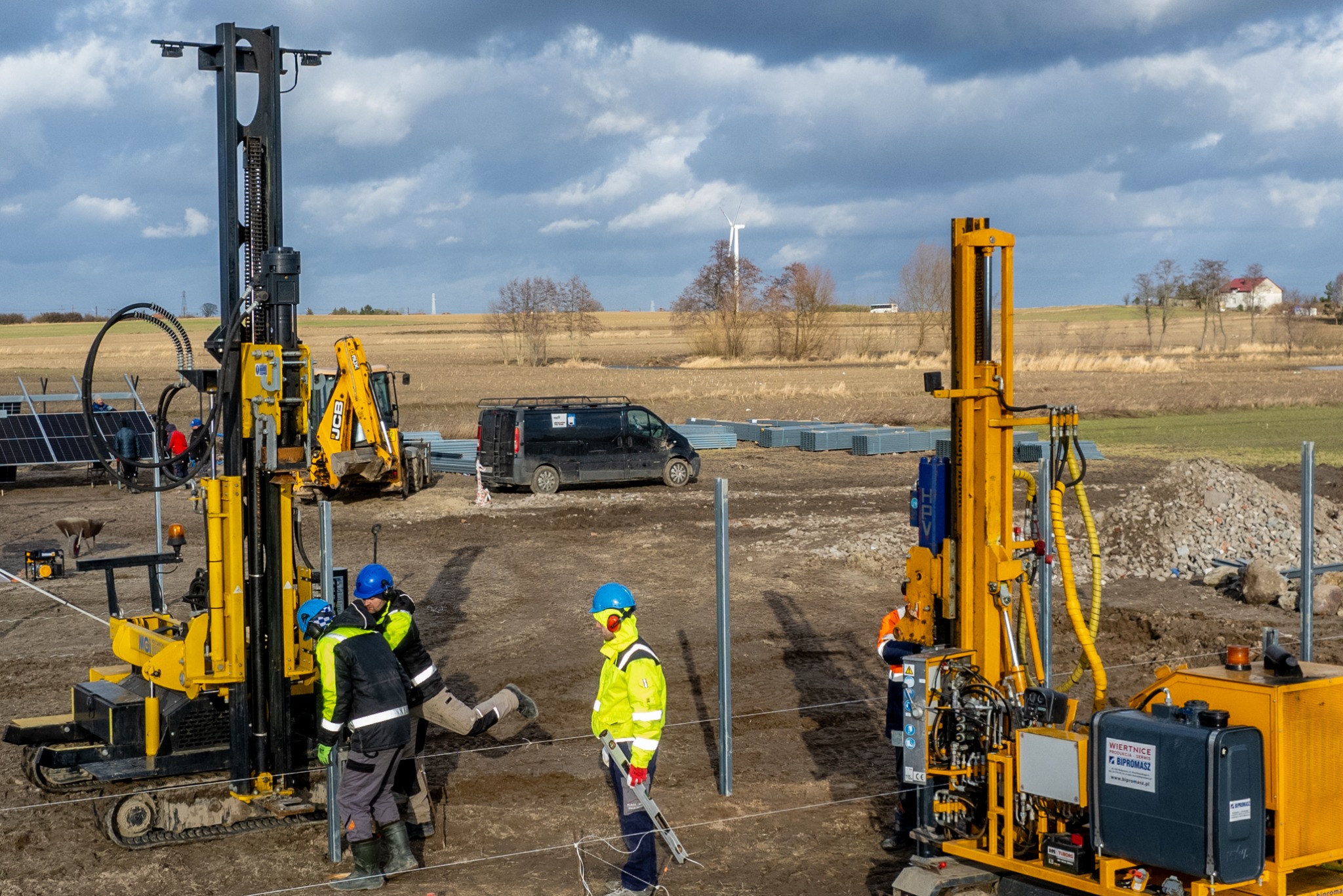 Prace ziemne pod instalację paneli: Dwóch pracowników w kaskach i odblaskowych kurtkach obsługuje żółtą wiertnicę na gąsienicach, w tle zaparkowany czarny van i widoczne elementy konstrukcji...
