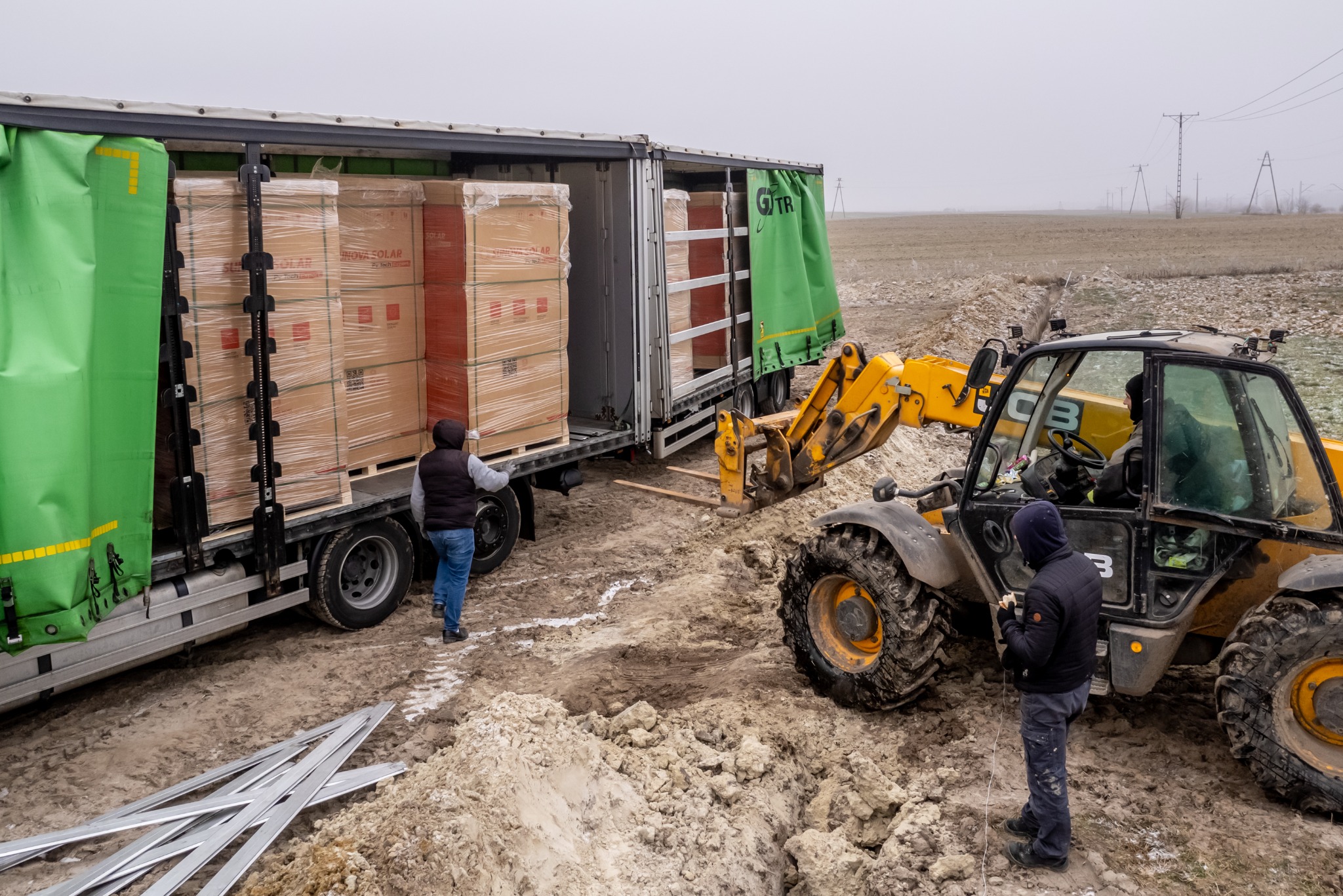 Rozładunek palet z panelami słonecznymi z ciężarówki za pomocą wózka widłowego JCB na placu budowy; widoczne elementy montażowe paneli.