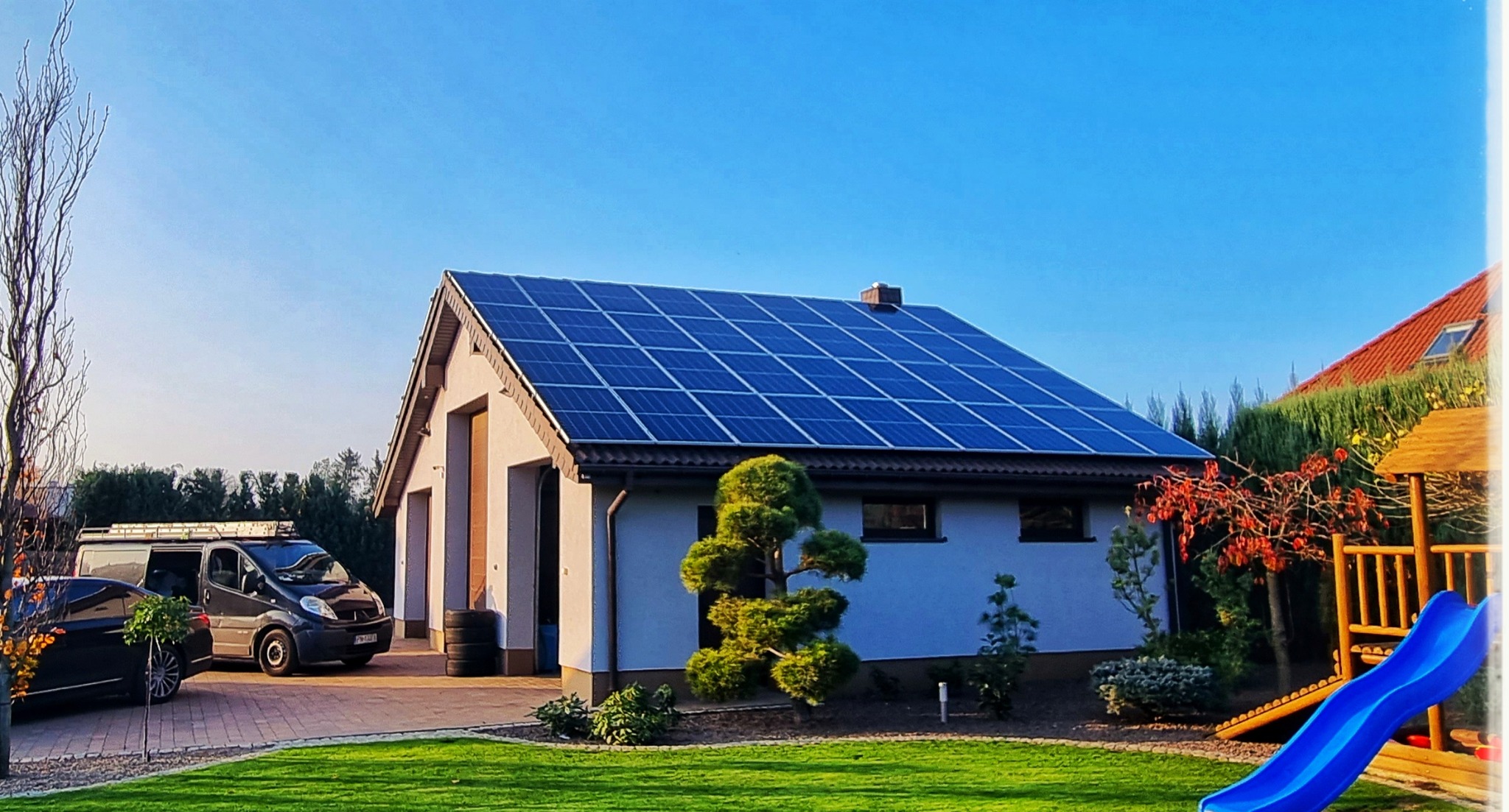Dom jednorodzinny z panelami słonecznymi na dachu, zaparkowany samochód dostawczy z drabiną na dachu, zadbany trawnik i ozdobne drzewko bonsai na pierwszym planie.