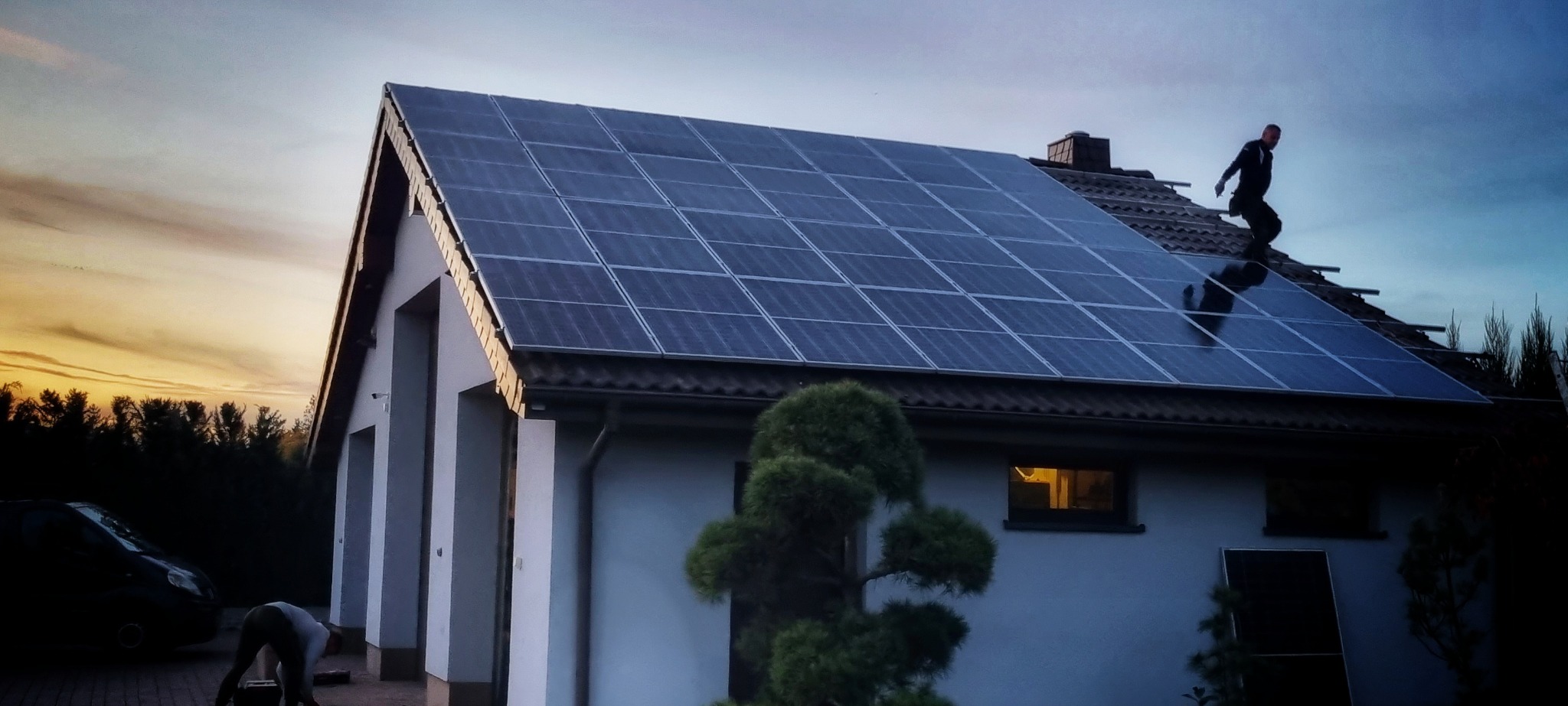 Montaż paneli fotowoltaicznych na dachu domu jednorodzinnego o zmierzchu, widoczny pracownik w kombinezonie na dachu i osoba pracująca na ziemi, częściowo widoczny samochód.