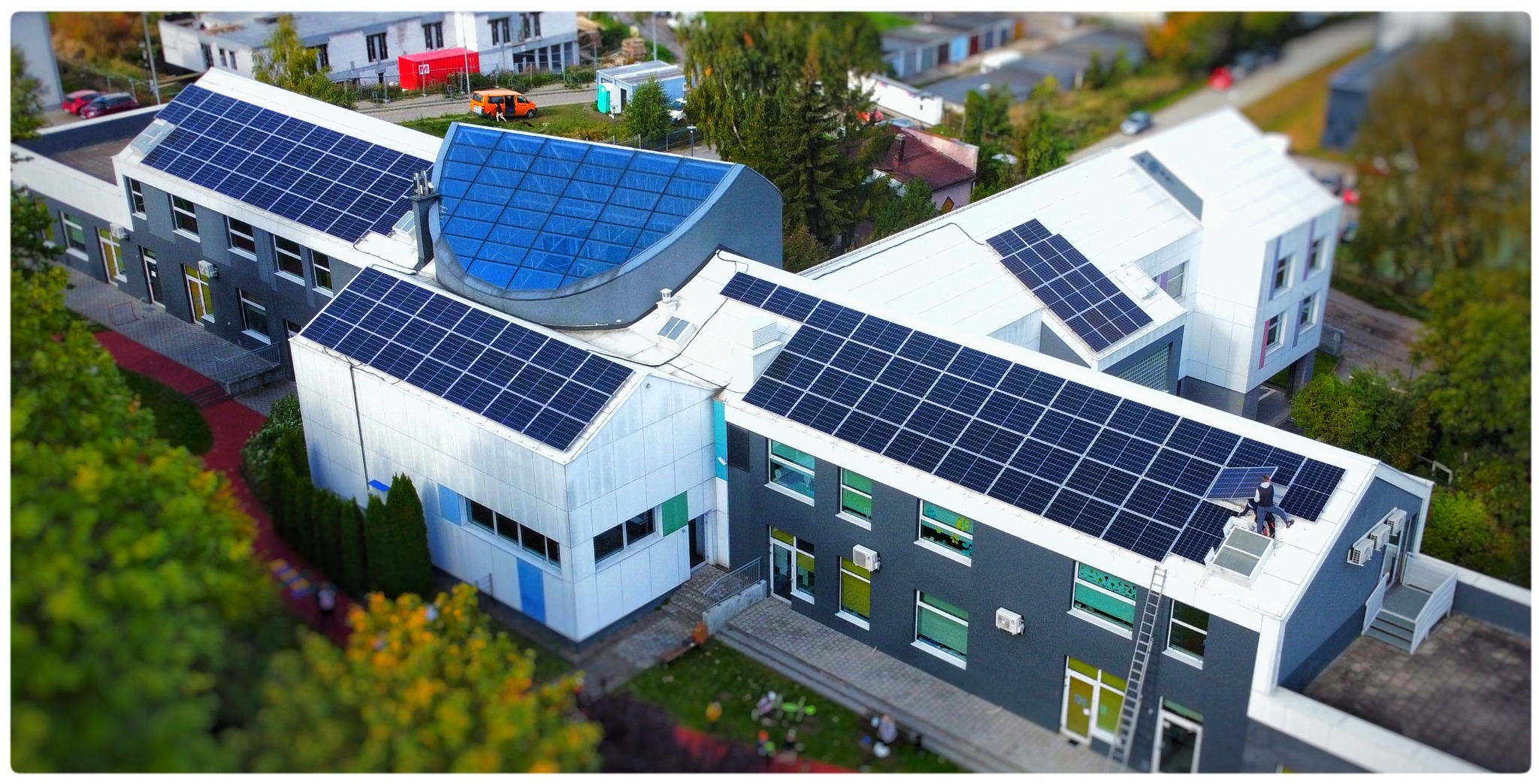 Widok z góry na budynek z zainstalowanymi panelami słonecznymi na dachu, gdzie jeden pracownik montuje dodatkowy panel, drabina oparta o ścianę budynku.