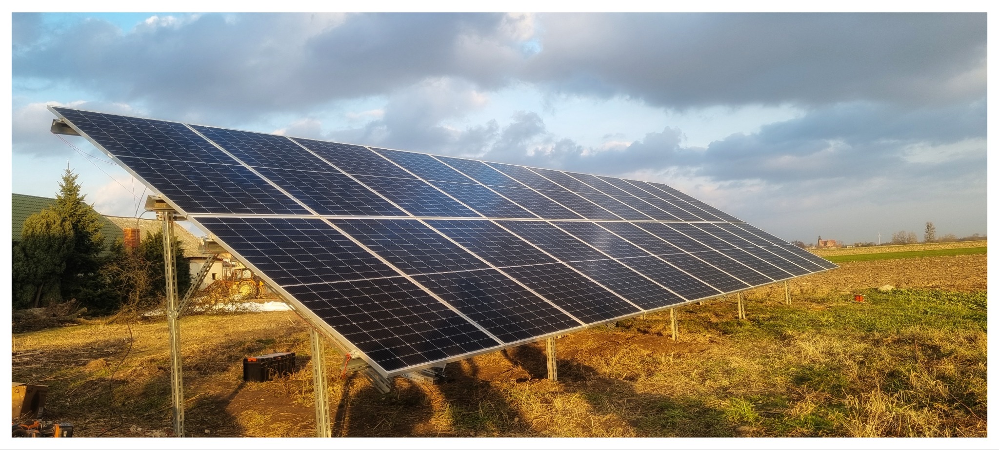 Duży panel słoneczny zamontowany na metalowej konstrukcji na polu, widoczny budynek mieszkalny i zachmurzone niebo w tle.