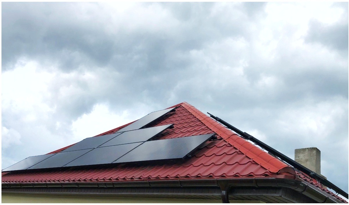 Część dachu pokrytego czerwoną blachodachówką z zamontowanymi panelami słonecznymi w układzie schodkowym, na tle pochmurnego nieba.