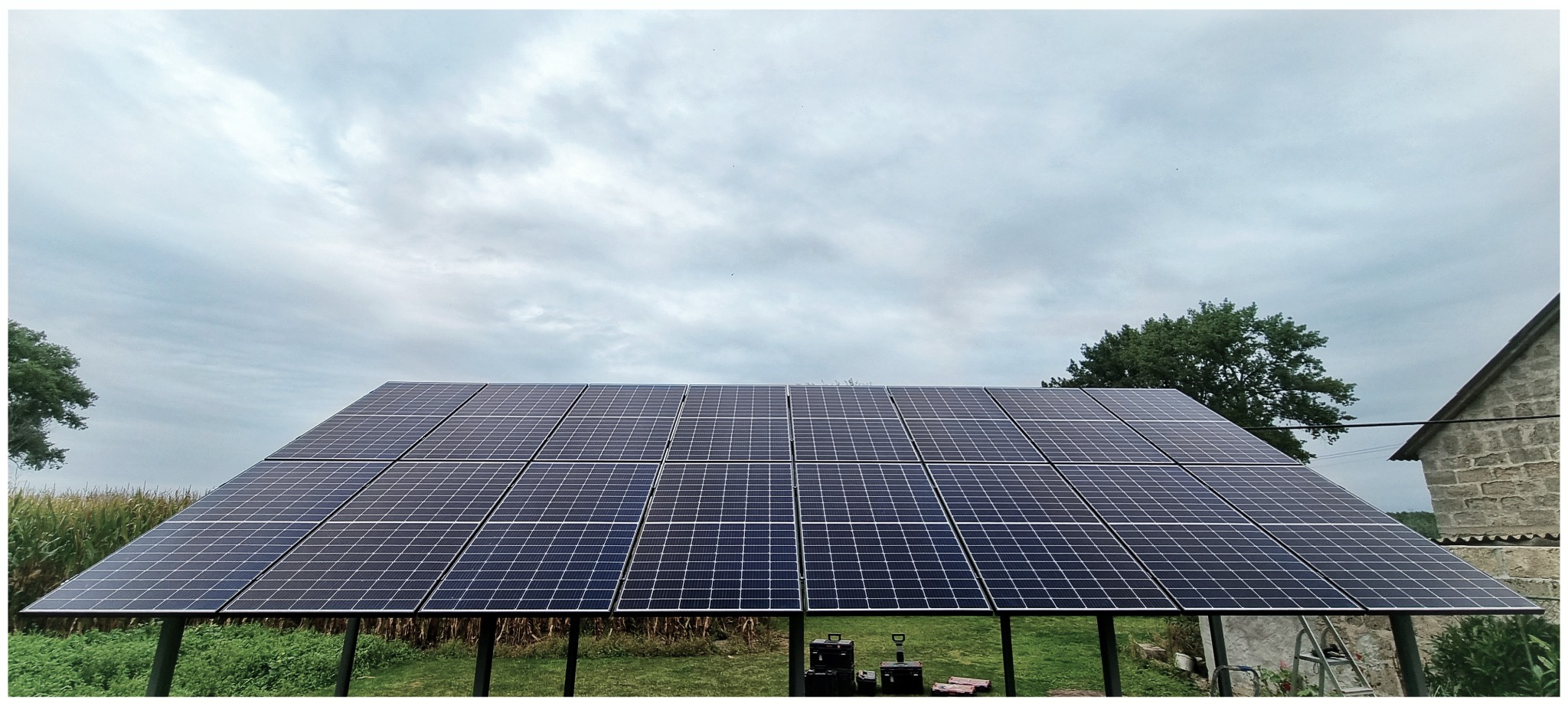 Zestaw paneli słonecznych na metalowej konstrukcji, zamontowany na trawiastym terenie z widokiem na pole kukurydzy i pochmurne niebo, obok budynku gospodarczego.