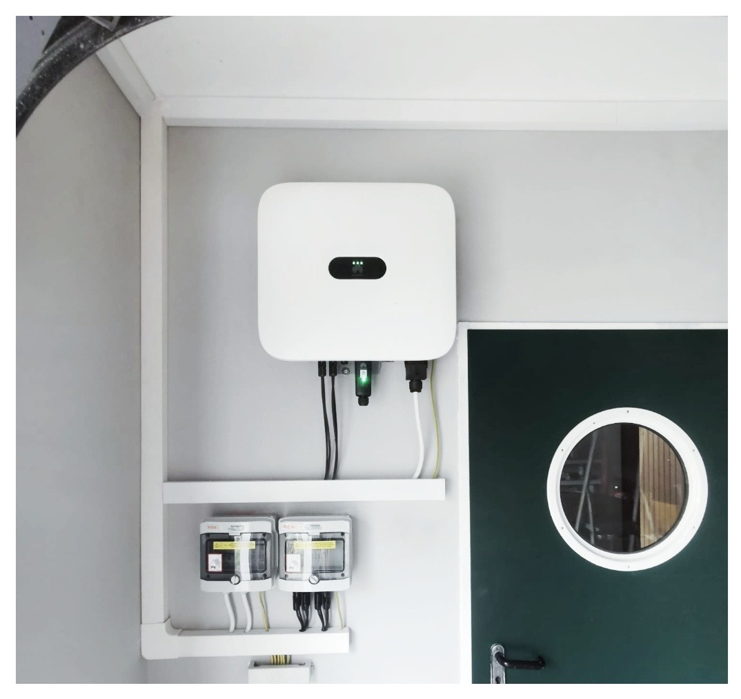 Biały inwerter fotowoltaiczny marki Huawei zamontowany na ścianie wewnątrz pomieszczenia, z widocznymi kablami i licznikami energii, obok zielonych drzwi z okrągłym okienkiem.