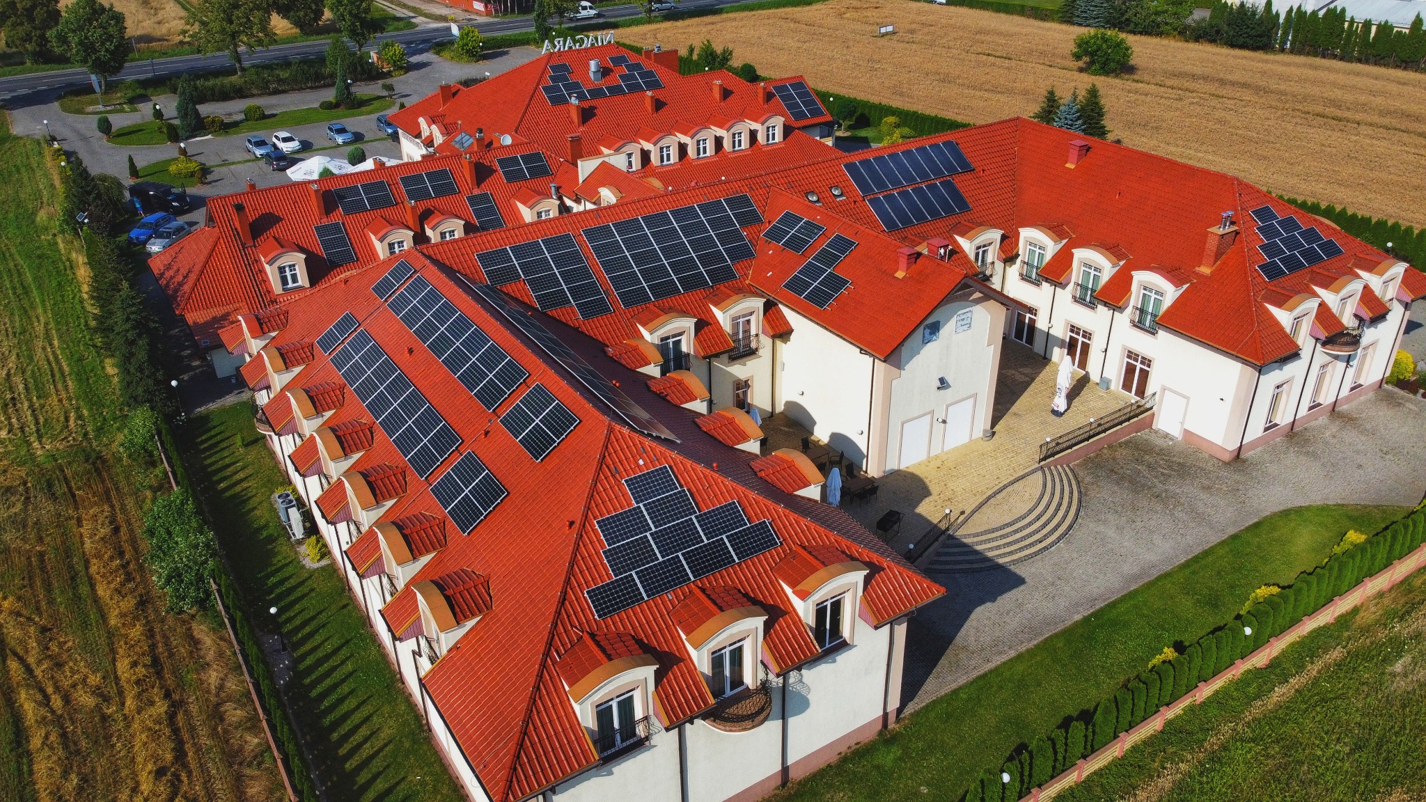 Widok z lotu ptaka na duży budynek z czerwonym dachem pokrytym panelami słonecznymi, otoczony zielenią i parkingiem.