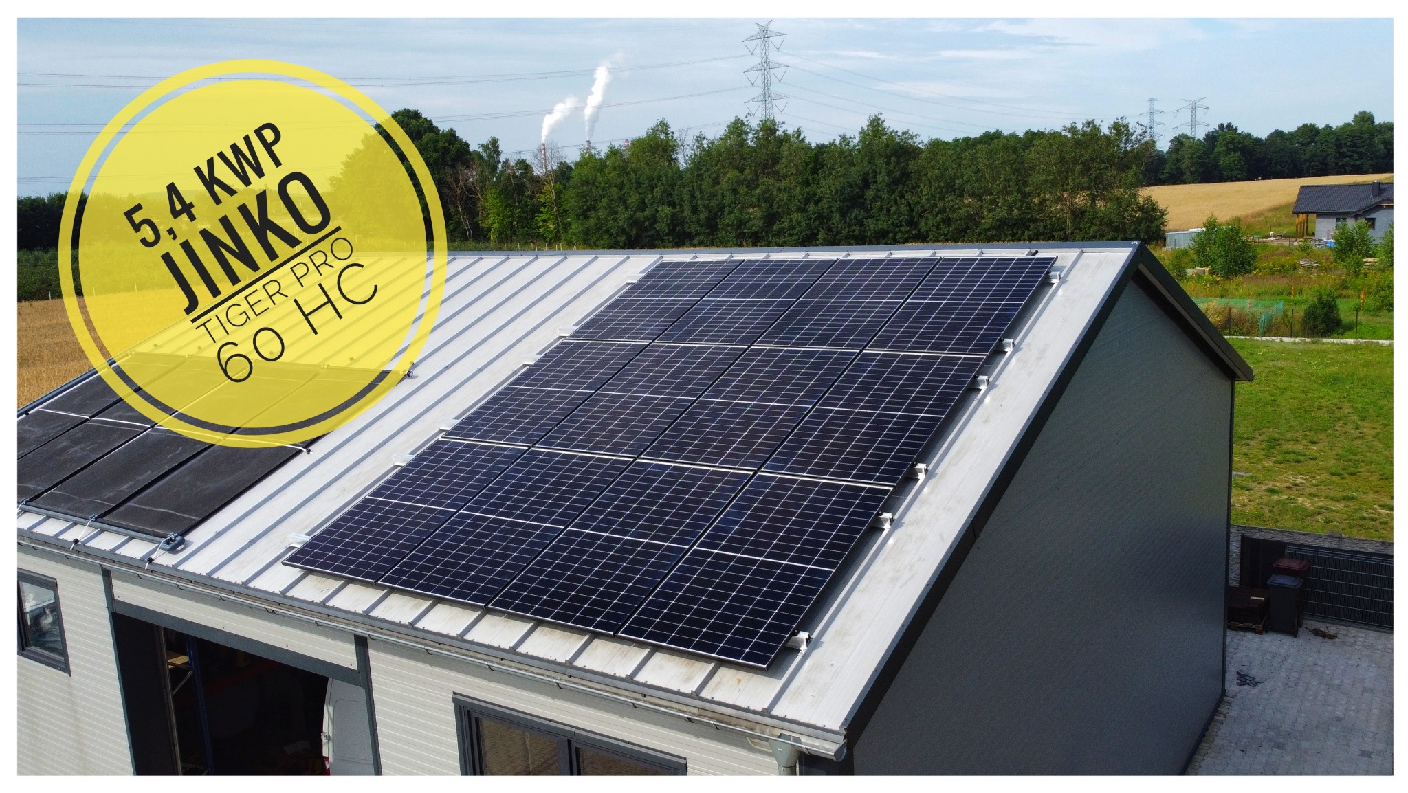 Panel słoneczny Jinko Tiger Pro 60 HC o mocy 5.4 kWp zamontowany na metalowym dachu budynku gospodarczego, w tle słupy wysokiego napięcia i komin