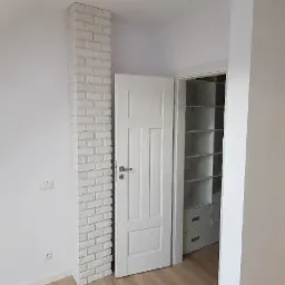 Wnętrze pokoju z kolumną z białej cegły dekoracyjnej, białe drzwi otwarte na białą garderobę z półkami i szufladami, jasne ściany i drewniana podłoga.