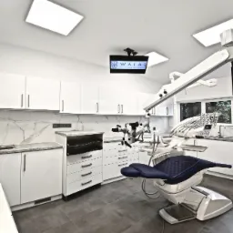 Nowoczesny gabinet stomatologiczny z niebieskim fotelem pacjenta, mikroskopem zabiegowym i białymi szafkami z marmurowym blatem, oświetlony lampami LED. Na ścianie widoczny monitor z logo kliniki...