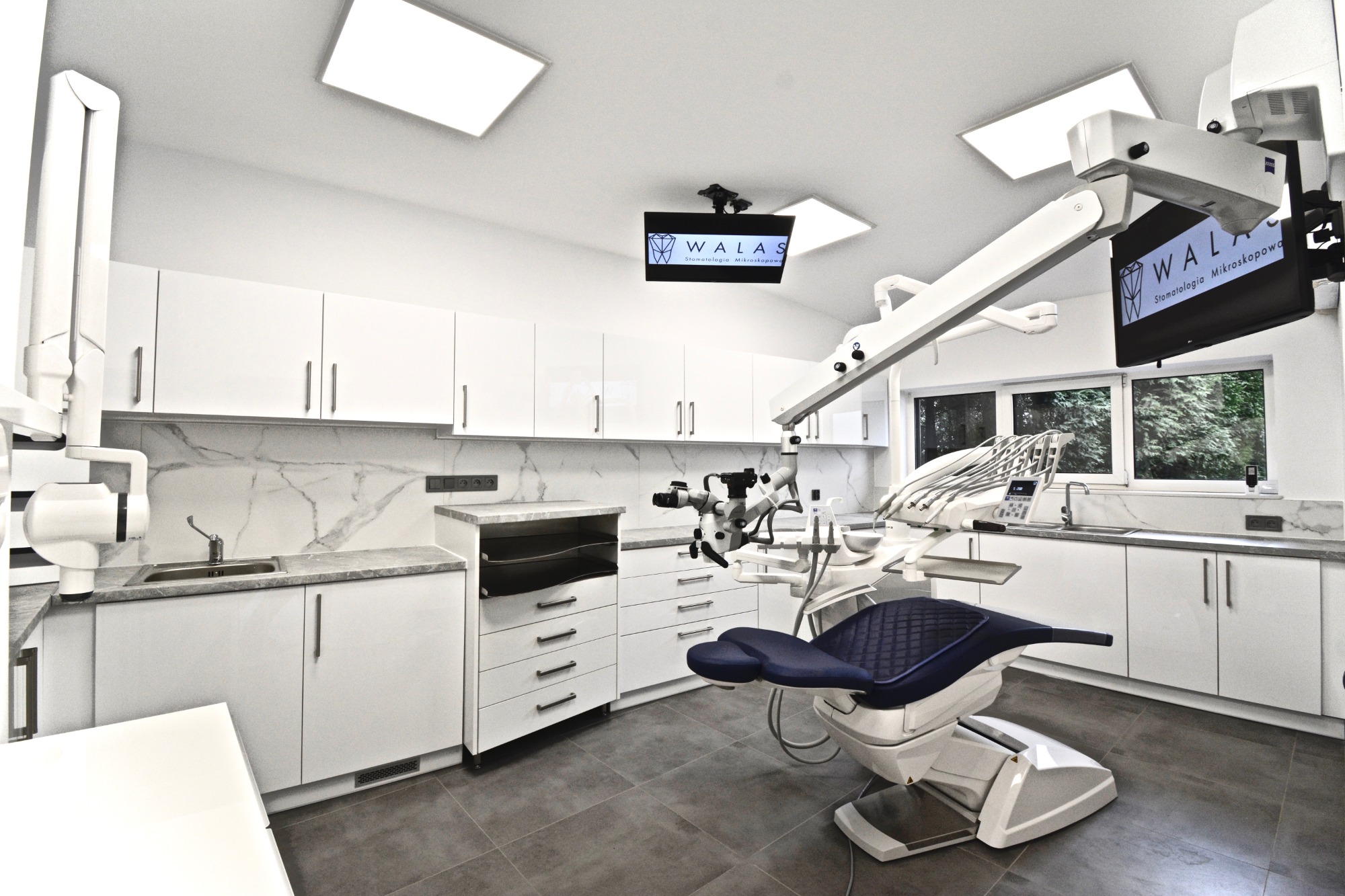 Nowoczesny gabinet stomatologiczny z niebieskim fotelem pacjenta, mikroskopem zabiegowym i białymi szafkami z marmurowym blatem, oświetlony lampami LED. Na ścianie widoczny monitor z logo kliniki...