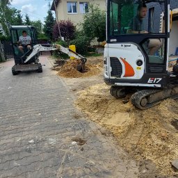 Kop Wiktor Gniado - Dwa minikoparki Bobcat na brukowanej drodze, jedna z operatorem w kabinie, druga w trakcie pracy z wysięgnikiem i łyżką, z usypaną pryzmą piasku.