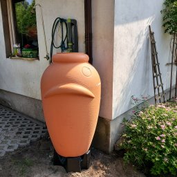 Kop Wiktor Gniado - Pomarańczowy zbiornik na deszczówkę w kształcie amfory, ustawiony przy białej ścianie domu, z podłączonym wężem i kranem.