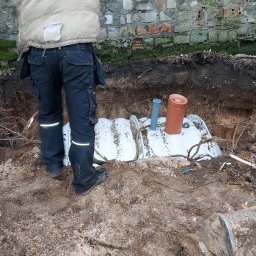 Kop Wiktor Gniado - Pracownik w odzieży roboczej montuje zbiornik przydomowej oczyszczalni ścieków w wykopie, widoczny fragment ściany z cegły w tle.
