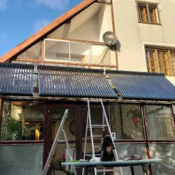 Panele solarne zamontowane nad przeszkloną werandą domu jednorodzinnego, widoczne drabiny i st&oacute;ł z narzędziami, błękitne niebo w tle.