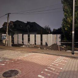 Kop Wiktor Gniado - Szare betonowe prefabrykaty ustawione wzdłuż chodnika z kostki, zabezpieczone taśmą ostrzegawczą, widoczny dom z drabina na dachu w tle, nocne oświetlenie.