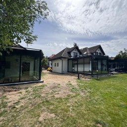 Kop Wiktor Gniado - Nowoczesna szklana zabudowa tarasowa z czarnymi ramami, widoczna na tle zadbanego ogrodu i domu z dwuspadowym dachem, w trakcie montażu, z piaskiem wokół konstrukcji i minikoparką w tle.