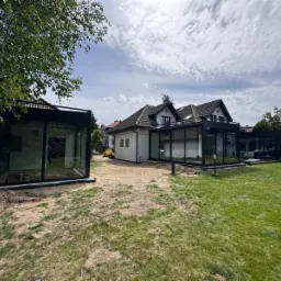 Nowoczesna szklana zabudowa tarasowa z czarnymi ramami, widoczna na tle zadbanego ogrodu i domu z dwuspadowym dachem, w trakcie montażu, z piaskiem wokół konstrukcji i minikoparką w tle.