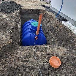 Kop Wiktor Gniado - Niebieski zbiornik z zieloną pokrywą umieszczony w wykopie ziemnym obok budynku mieszkalnego, widoczny pomarańczowy przewód i pokrywa na ziemi.