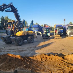 Kop Wiktor Gniado - Żółta koparka Mecalac MXT z operatorem w kabinie, na placu budowy z hałdą ziemi w tle, obok widoczne inne maszyny budowlane i ciężarówki.