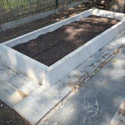 Kop Wiktor Gniado - Podwyższona rabata z betonowych elementów, wypełniona ziemią, otoczona kostką brukową. W tle widoczne ogrodzenie i drzewo. Całość w słoneczny dzień.