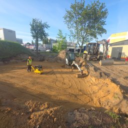 Kop Wiktor Gniado - Niwelacja terenu z użyciem zagęszczarki i minikoparki na placu budowy. Pracownik w kamizelce odblaskowej. W tle budynki i drzewa.