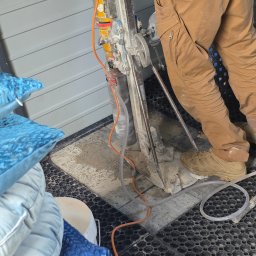 Kop Wiktor Gniado - Pracownik wierci otwór w posadzce wiertnicą rdzeniową, widoczne przewody i woda chłodząca. W tle ściana z szarego sidingu i stos poduszek dekoracyjnych.