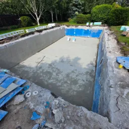 Etap budowy betonowego basenu w ogrodzie: wylany fundament, częściowo wyłożone ściany, widoczne elementy instalacji. Prace ziemne, gruz i narzędzia wokół.