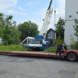 Kop Wiktor Gniado - Transport niskopodwoziowy z dźwigiem na tle zieleni i budynku. Widoczni dwaj mężczyźni przy naczepie. Dźwig w kolorach białym i niebieskim.