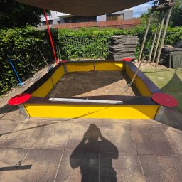 Kop Wiktor Gniado - Nowoczesna piaskownica z żółtymi ściankami i czerwoną obwódką na tle zielonego ogrodu, częściowo zacieniona. Widoczne narzędzia i materiały budowlane.