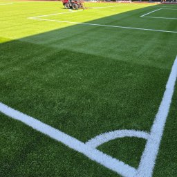 Kop Wiktor Gniado - Fragment boiska ze sztuczną trawą, wyznaczony białymi liniami, w tle maszyna do pielęgnacji murawy. Widok z góry na róg boiska, oświetlony częściowo słońcem.