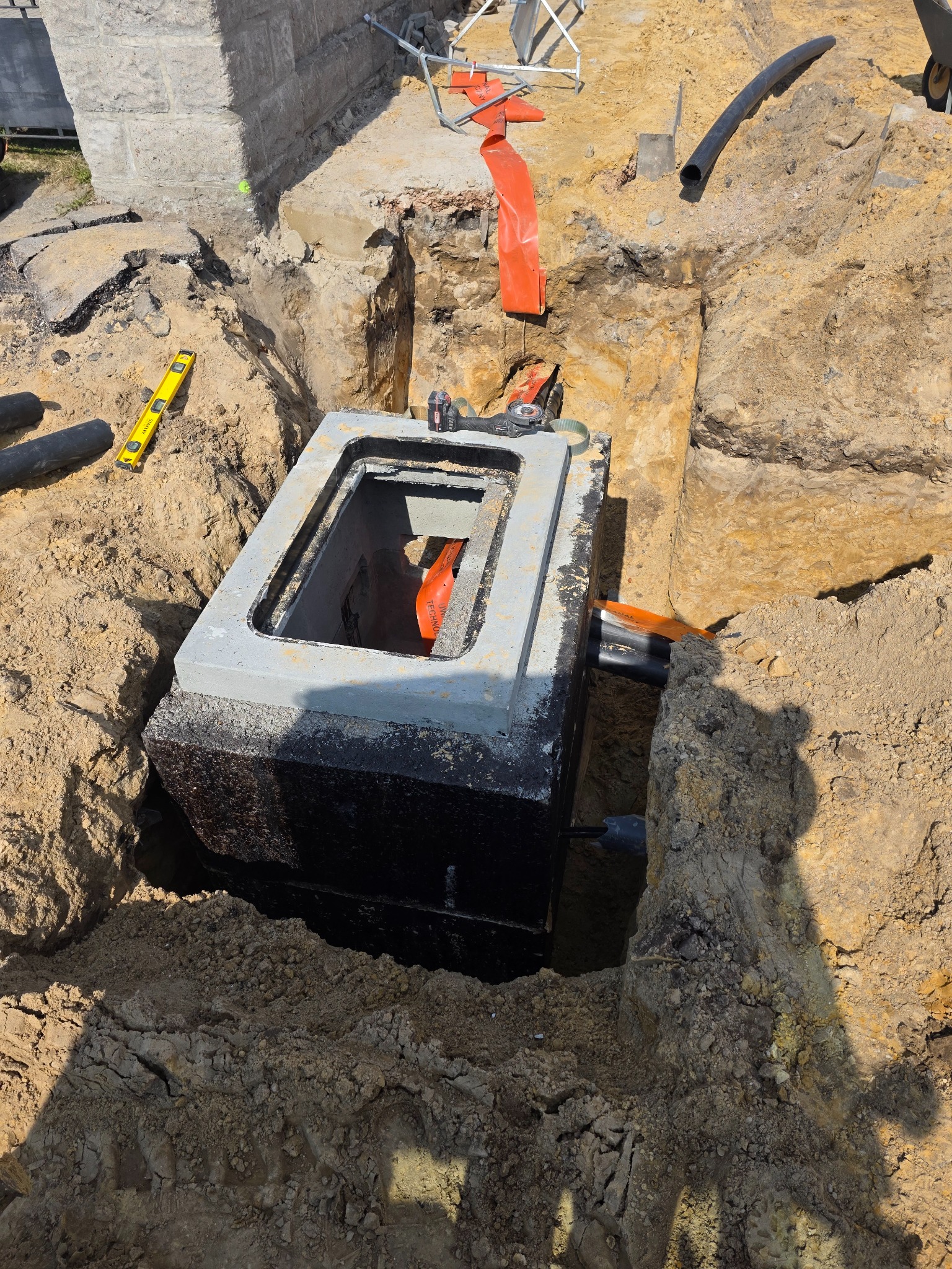 Betonowa studzienka kanalizacyjna w wykopie ziemnym, widoczne rury i narzędzia, poziomica Stanley i pomarańczowa taśma ostrzegawcza.