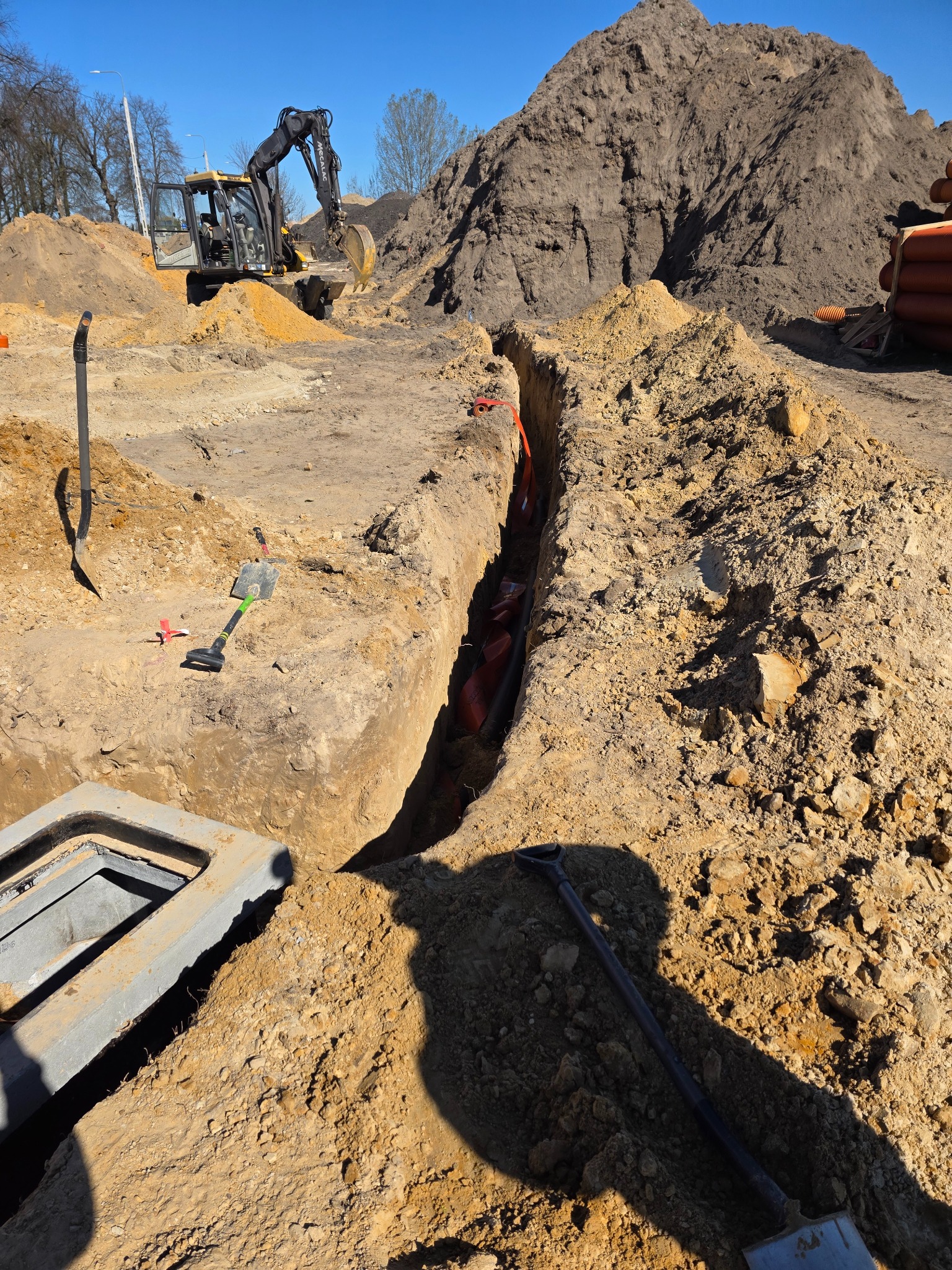 Wykop z rurami w ziemi, w tle koparka i sterta ziemi. Widoczne narzędzia: łopaty i szpadel. Otwarte, betonowe włazy kanalizacyjne. Słoneczny dzień.