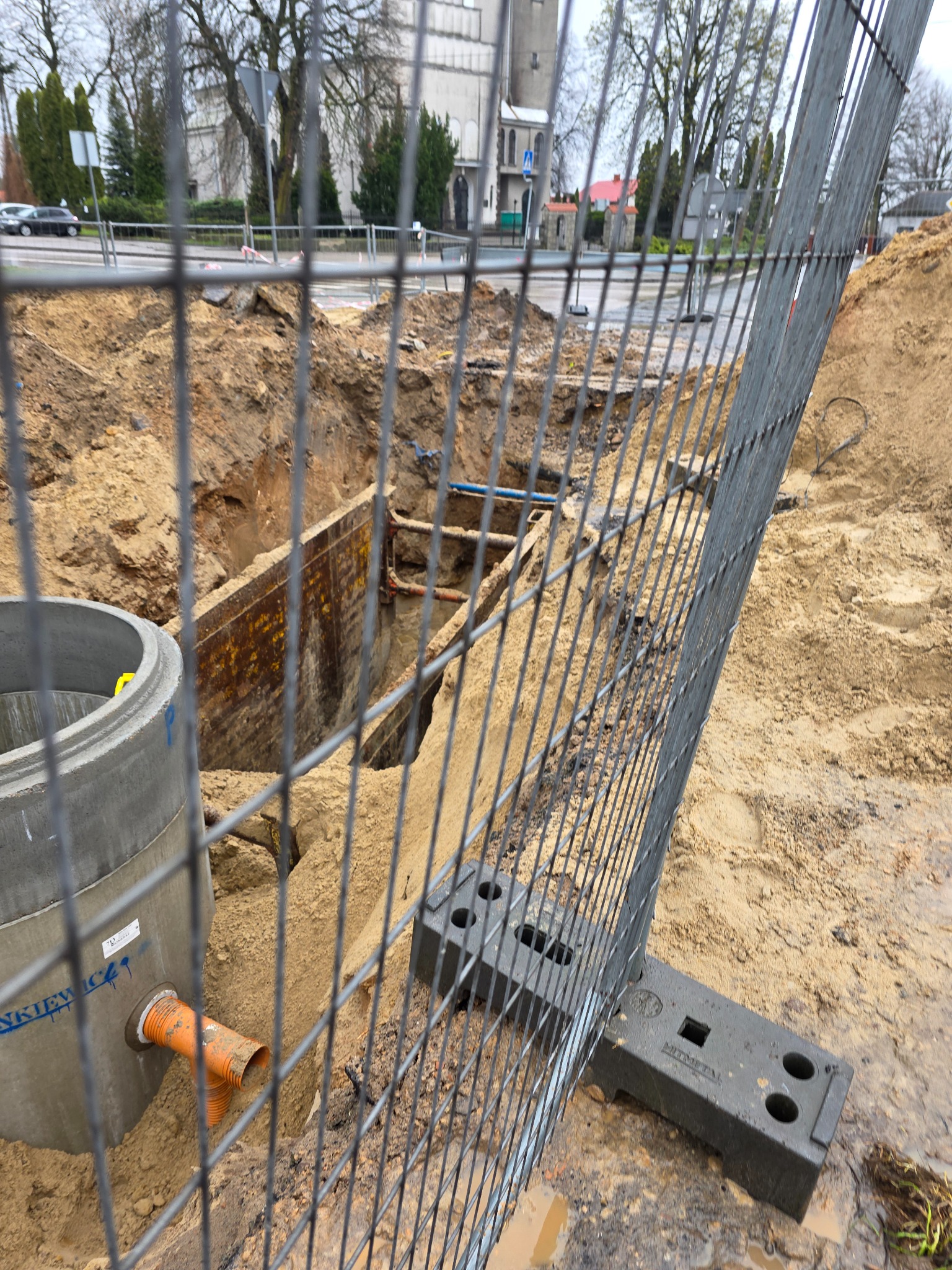 Wykop zabezpieczony siatką, widoczny betonowy krąg studni z pomarańczową rurą, w tle ziemia i budynek. Prace ziemne przy instalacji wod-kan.