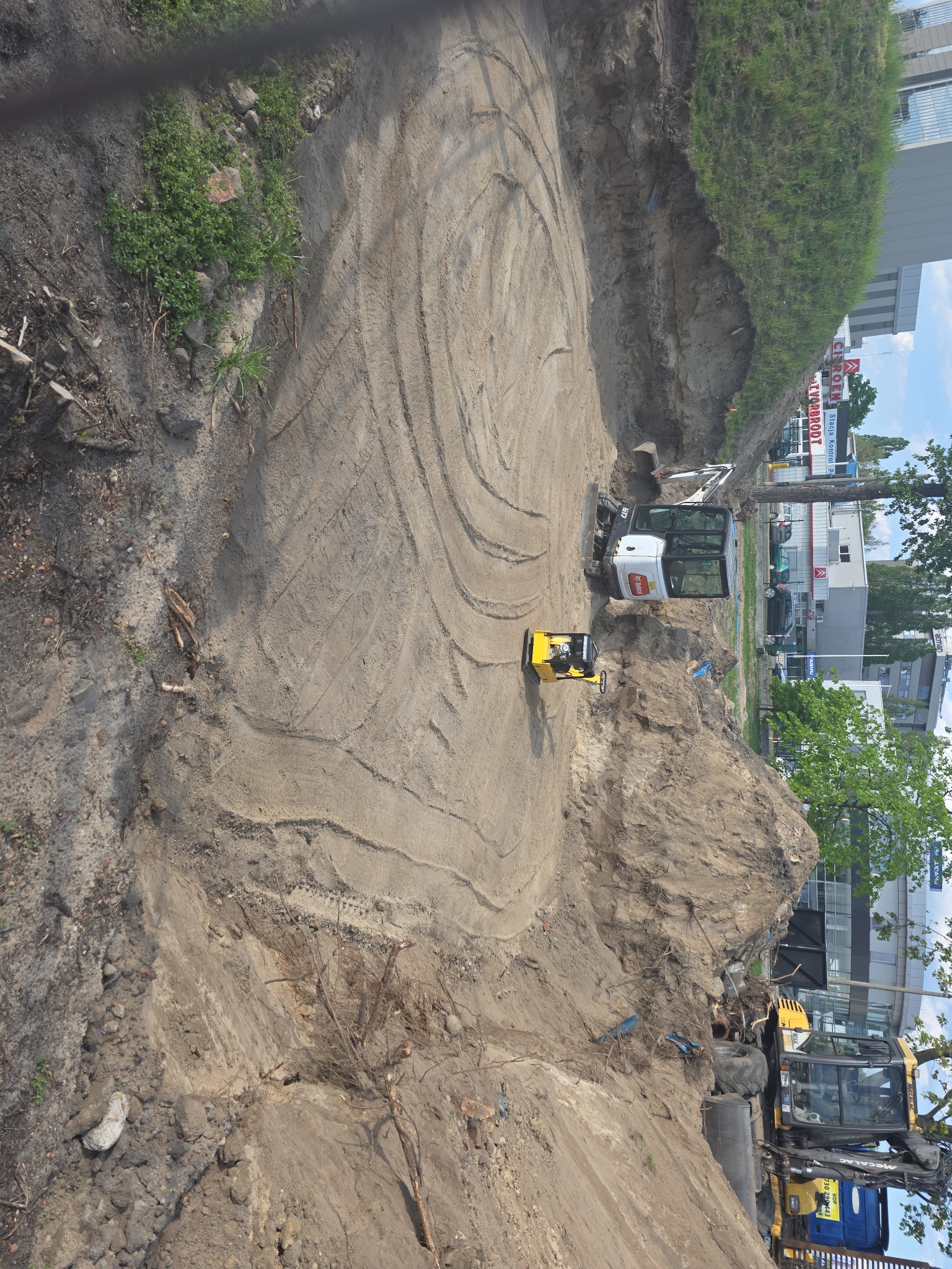 Żółty walec i biała mini koparka na placu budowy podczas niwelacji terenu. Widoczne ślady opon na piasku. Ujęcie z góry.