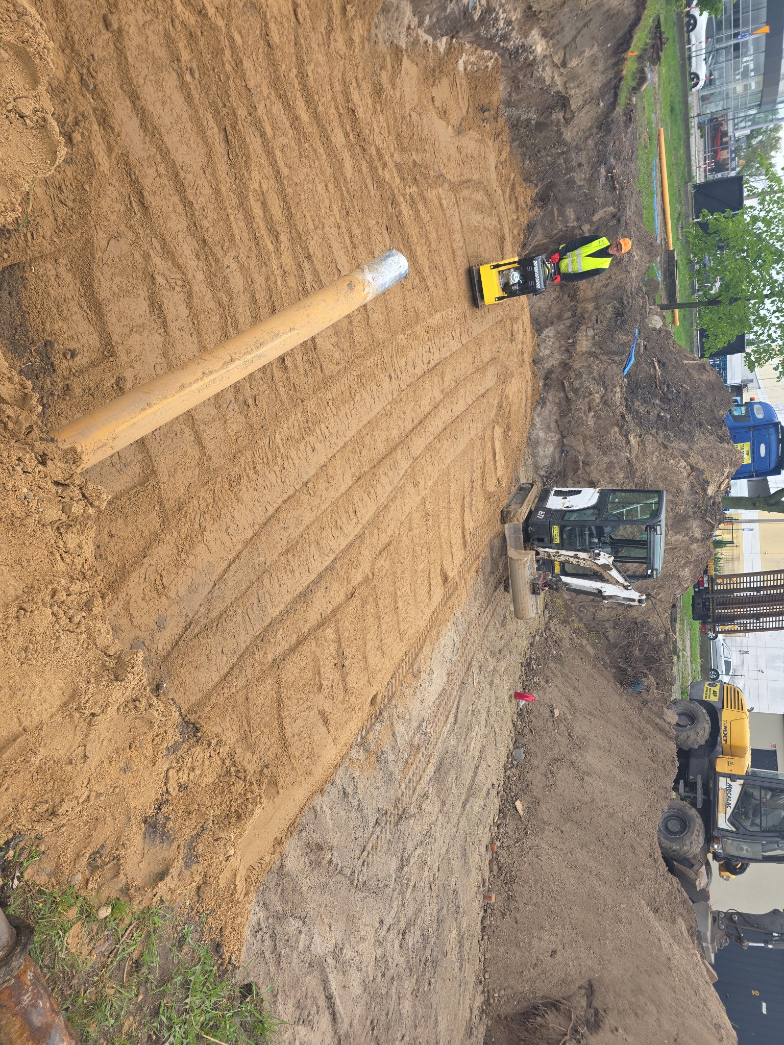 Żółty rurociąg w wykopie, ubijarka gruntu i mała koparka na placu budowy w słoneczny dzień. Widoczne ślady po przejeździe maszyn.
