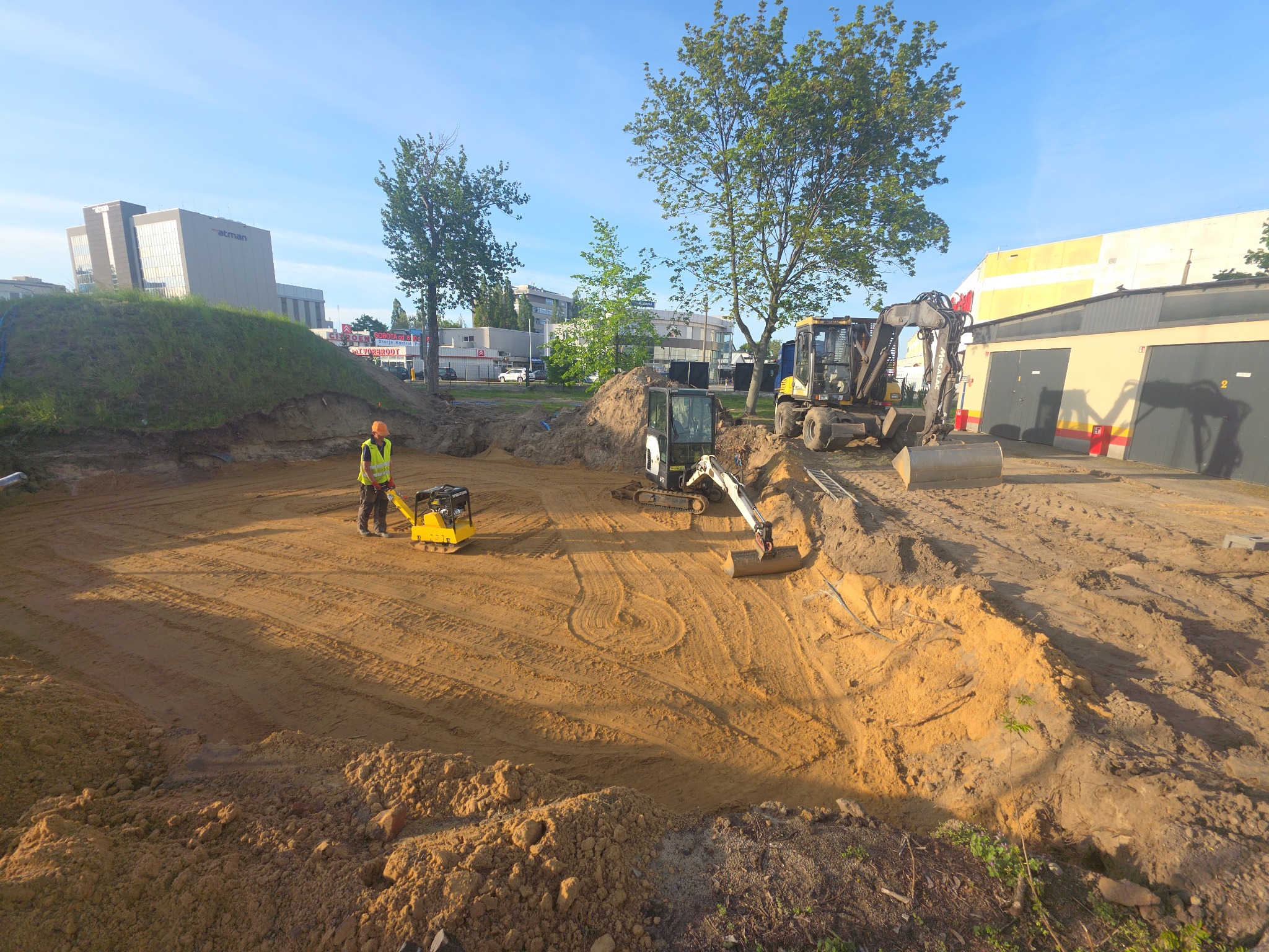 Niwelacja terenu z użyciem zagęszczarki i minikoparki na placu budowy. Pracownik w kamizelce odblaskowej. W tle budynki i drzewa.