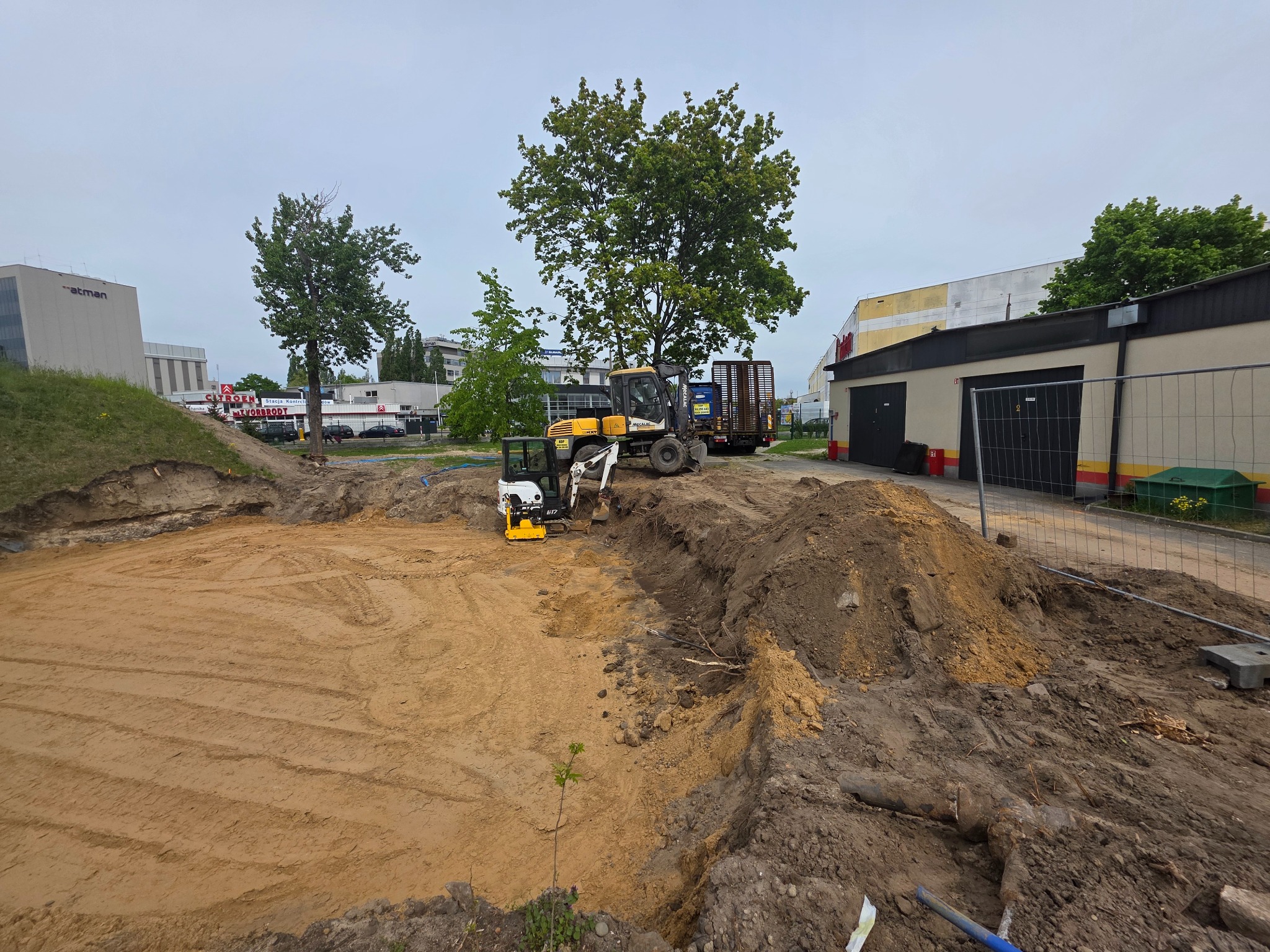 Żółty i biały mini-koparka na wykopie, przygotowany teren pod budowę z widocznymi śladami pracy maszyn, w tle budynki i drzewa.