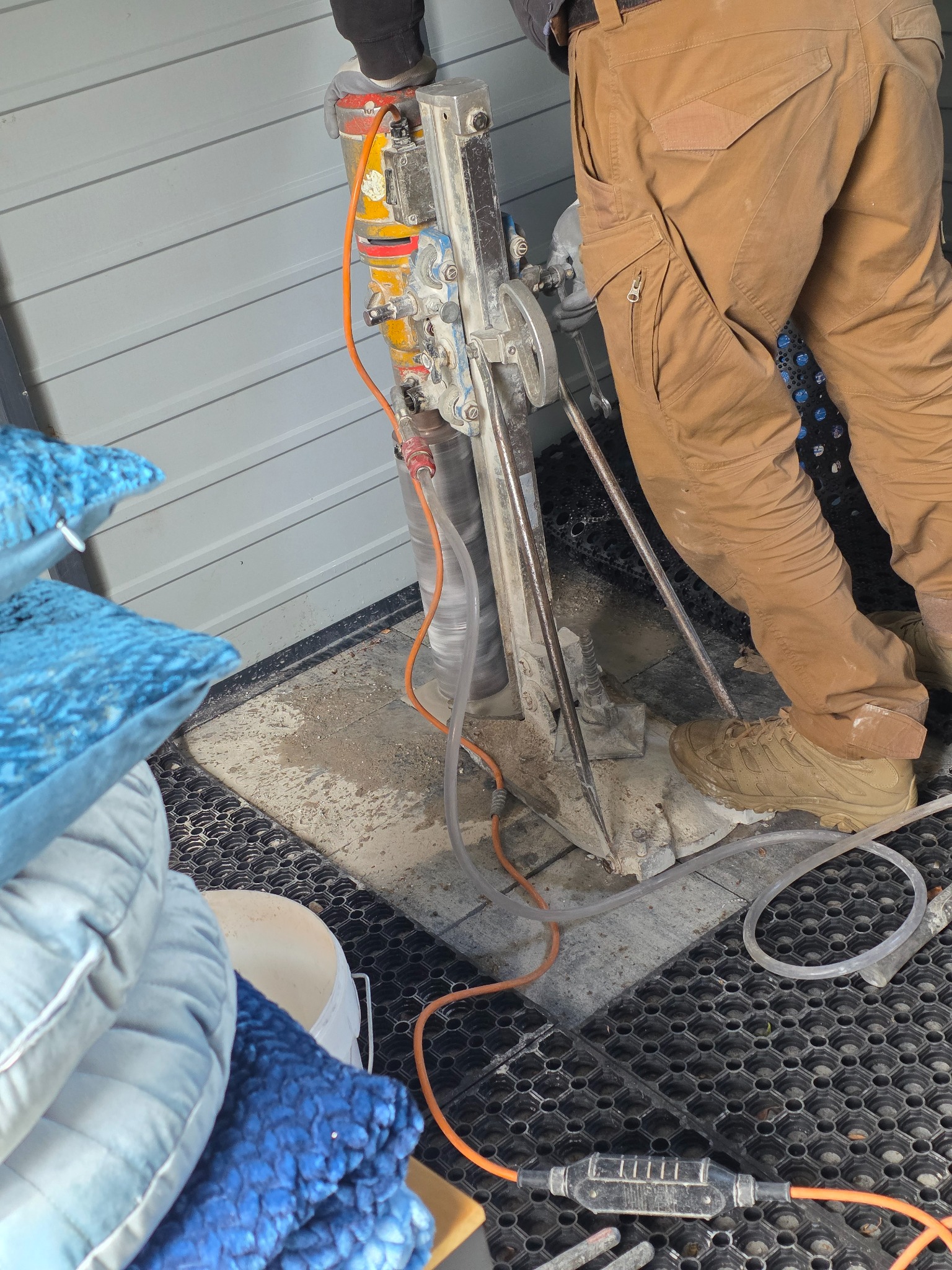 Pracownik wierci otwór w posadzce wiertnicą rdzeniową, widoczne przewody i woda chłodząca. W tle ściana z szarego sidingu i stos poduszek dekoracyjnych.