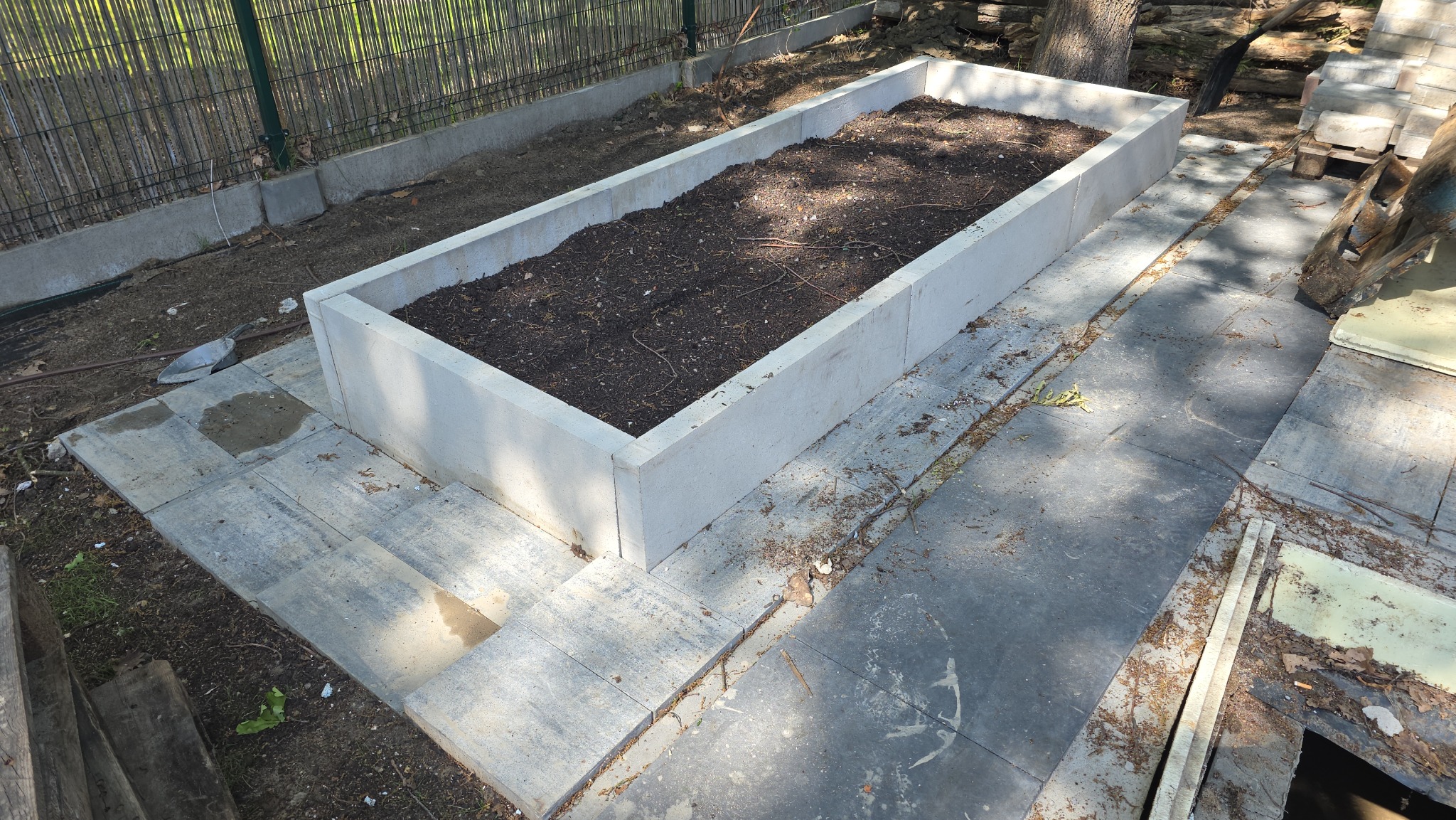Podwyższona grządka z betonowych elementów, wypełniona ziemią, otoczona kostką brukową i ogrodzeniem. Widoczna łopata i fragment pnia drzewa.