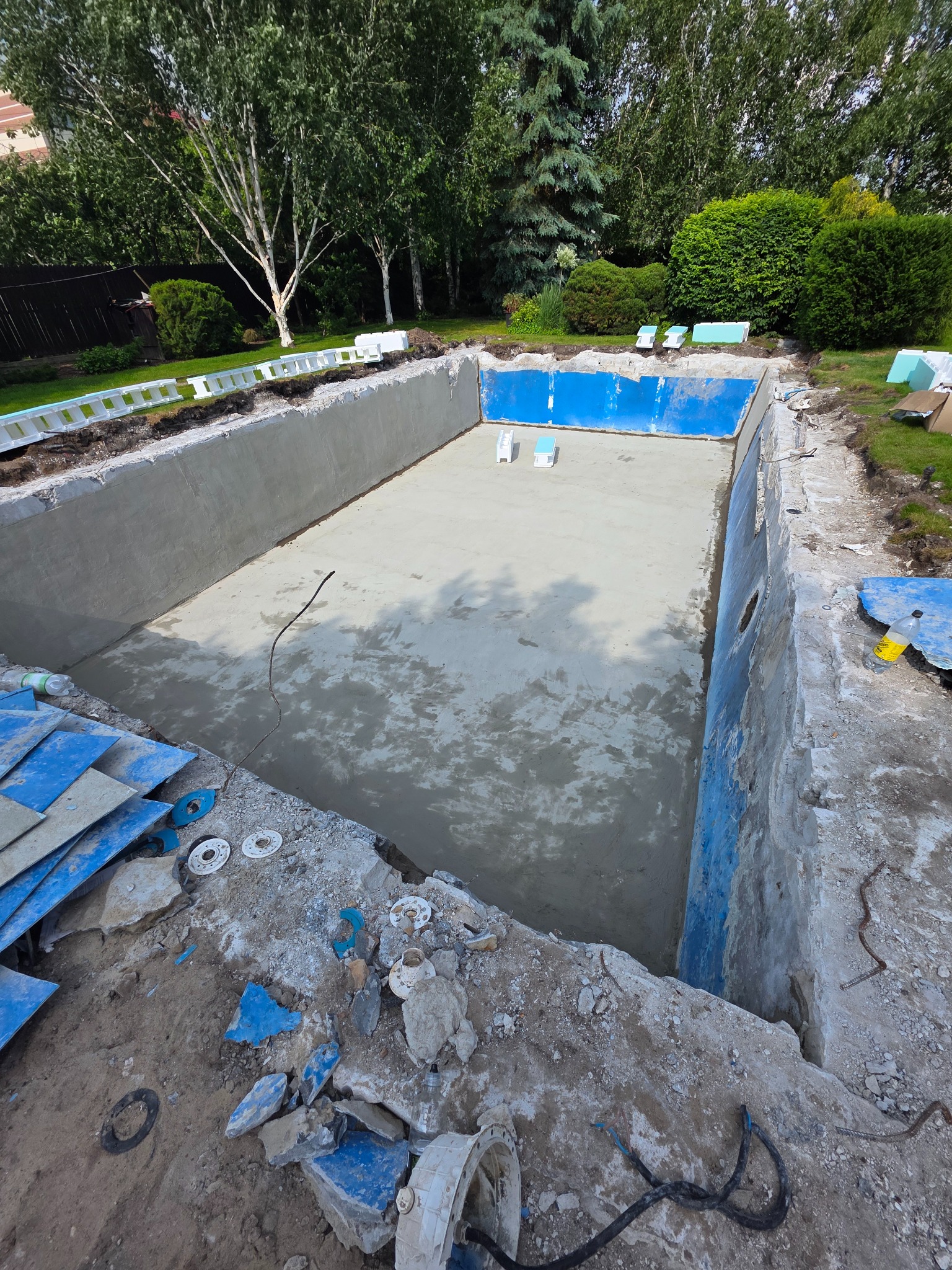 Etap budowy betonowego basenu w ogrodzie: wylany fundament, częściowo wyłożone ściany, widoczne elementy instalacji. Prace ziemne, gruz i narzędzia wokół.