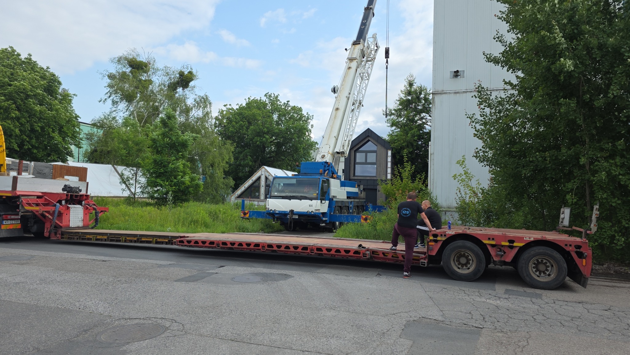 Transport niskopodwoziowy z dźwigiem na tle zieleni i budynku. Widoczni dwaj mężczyźni przy naczepie. Dźwig w kolorach białym i niebieskim.