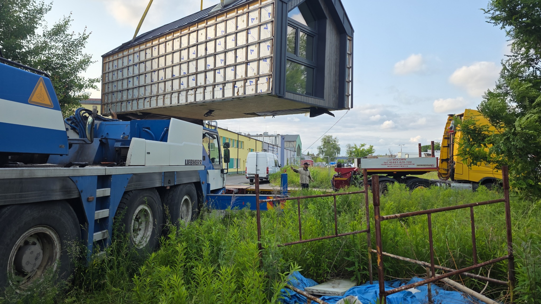Przenoszenie modułowego domu z widocznymi panelami izolacyjnymi za pomocą dźwigu Liebherr na tle zieleni i budynków. Transport domu na naczepie.