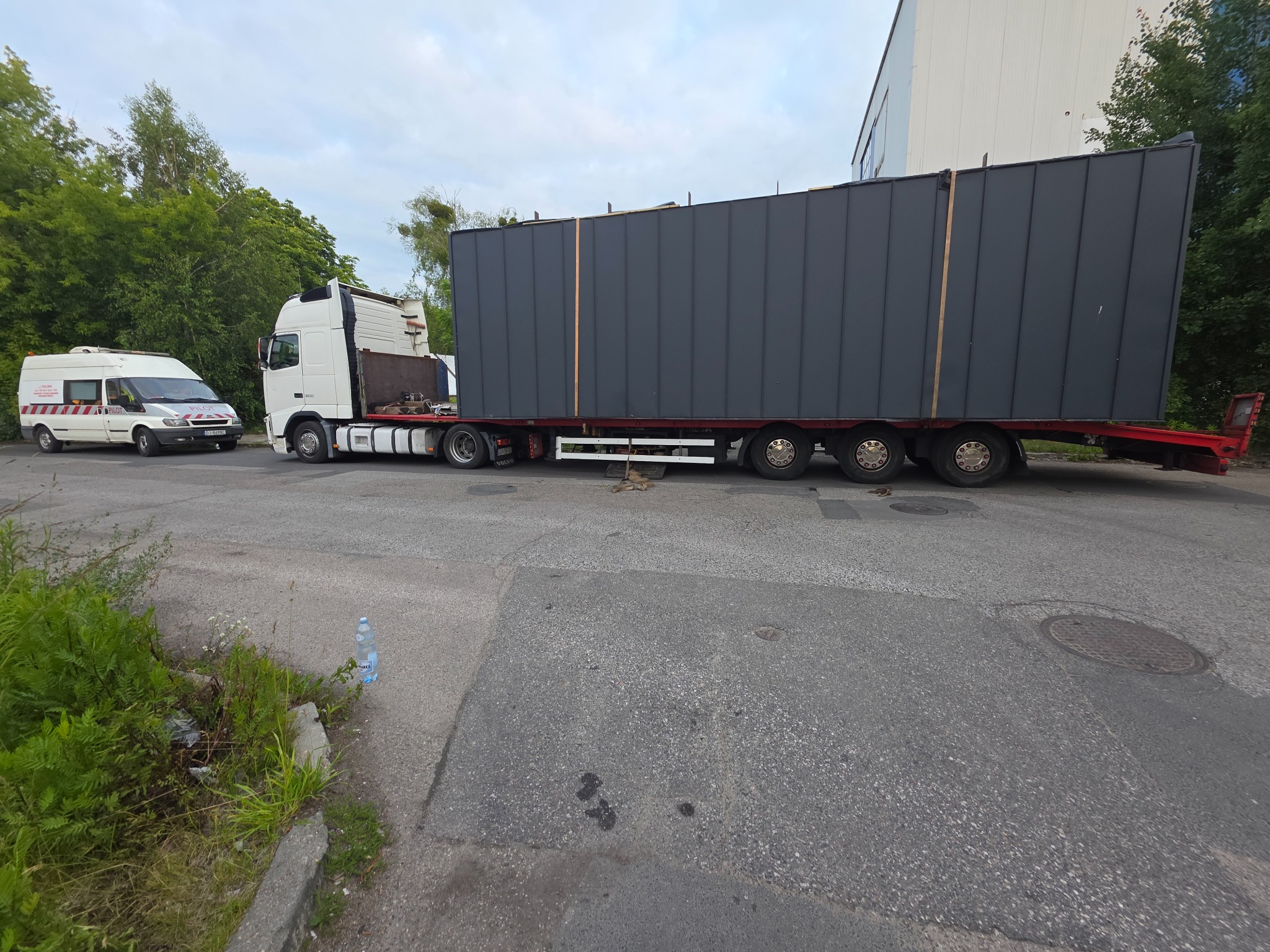 Transport niskopodwoziowy przewozi duży, szary moduł z pionowymi panelami. Biały van pilotujący i butelka wody przy krawężniku uzupełniają scenerię.