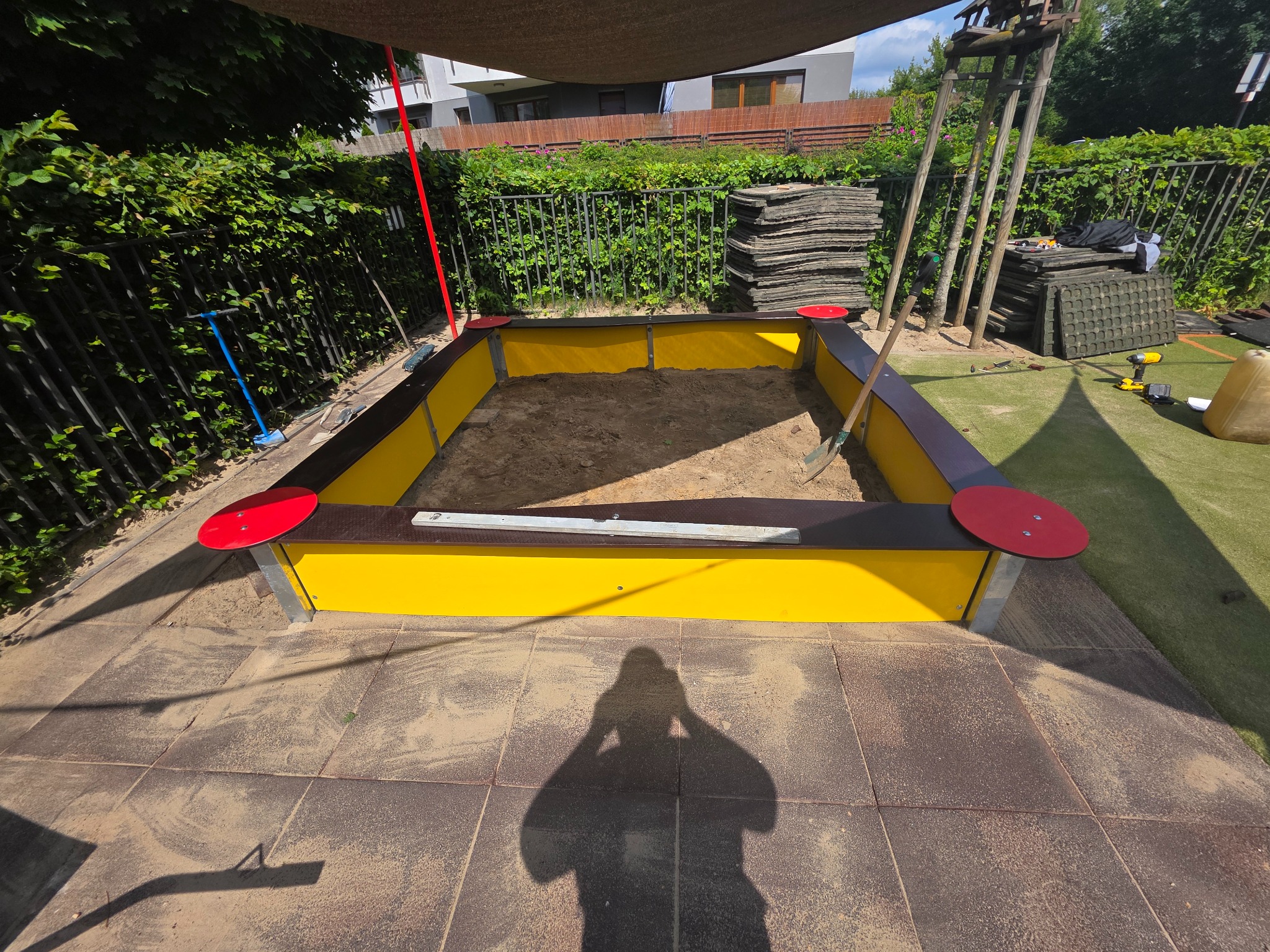 Nowoczesna piaskownica z żółtymi ściankami i czerwoną obwódką na tle zielonego ogrodu, częściowo zacieniona. Widoczne narzędzia i materiały budowlane.