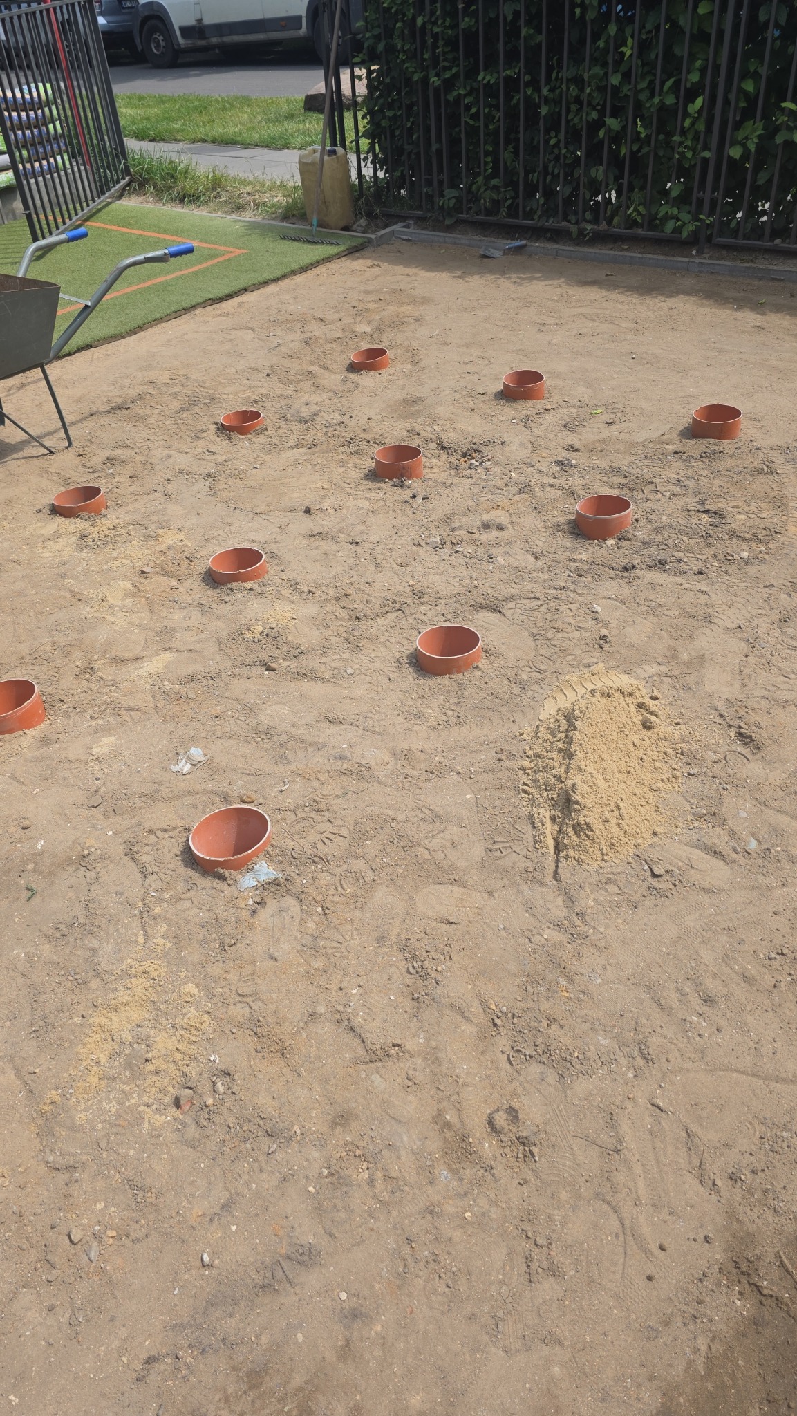 Przygotowanie terenu pod trawnik: rozmieszczone rury nawadniające na piasku, widoczna taczka i fragment trawnika w tle. Etap prac ziemnych.